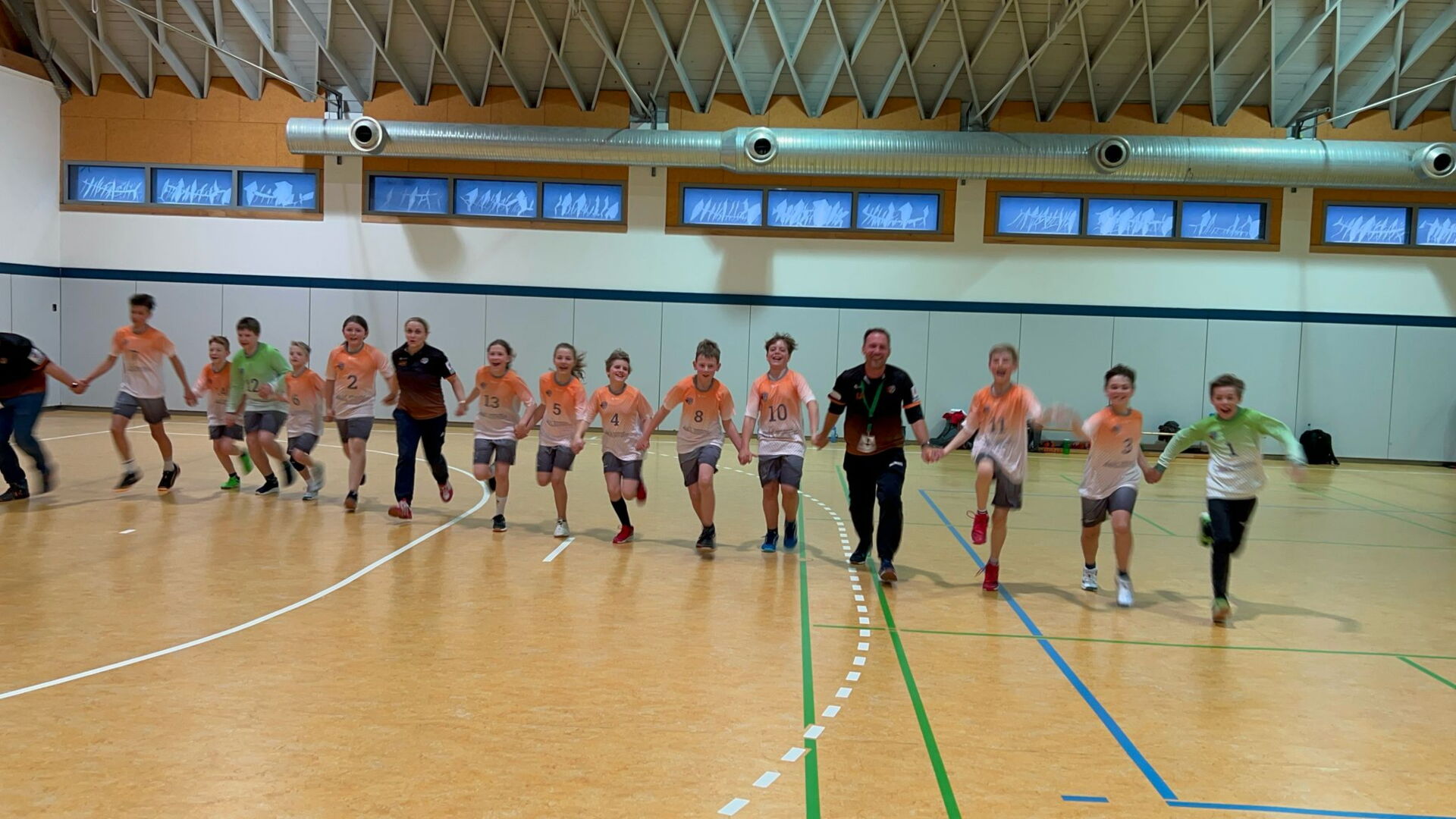 Eine Reihe junger Handballspieler der HSG Coswig, angeführt von einem Trainer, laufen Hand in Hand in einer Sporthalle.
