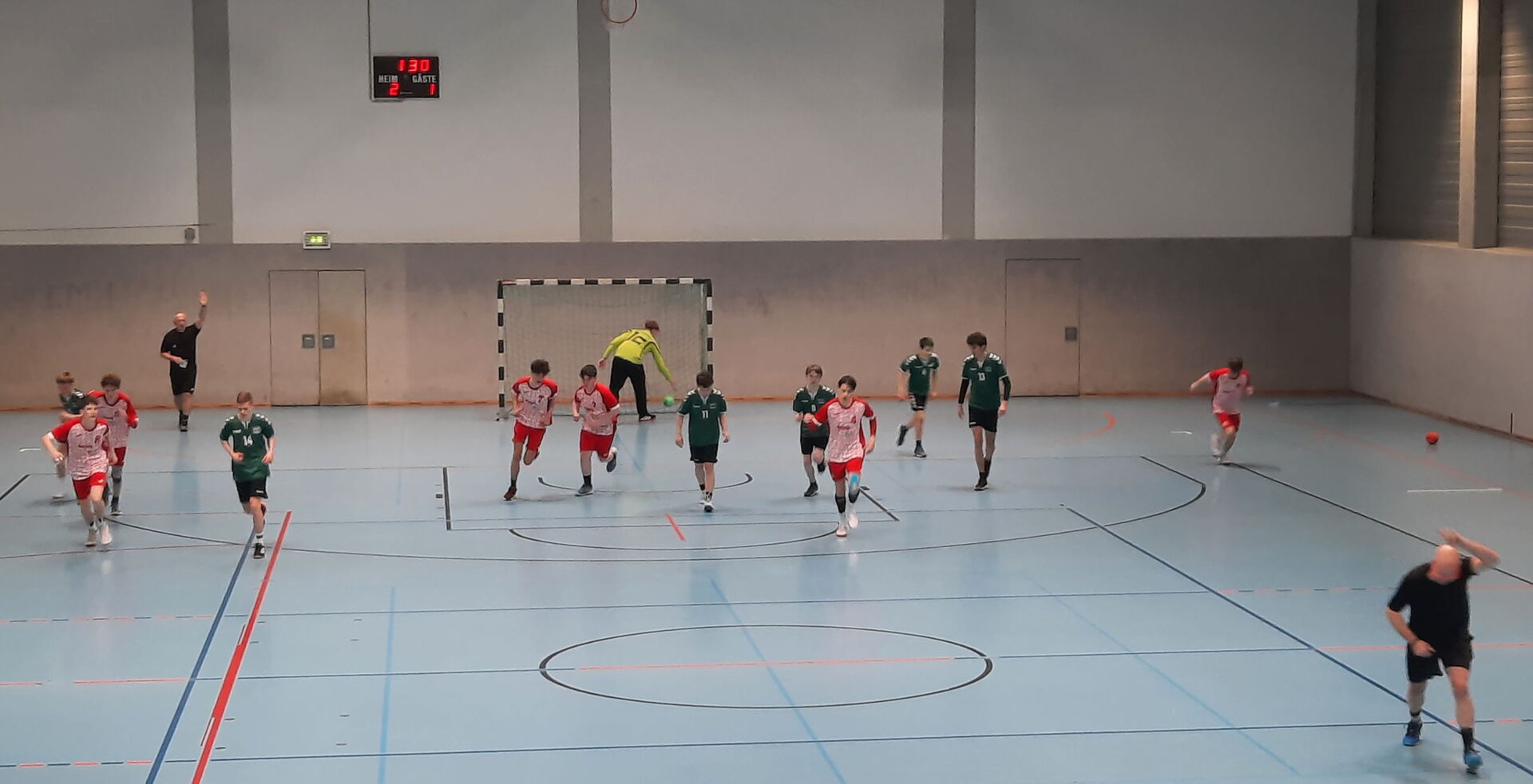 Eine Aufnahme aus der Vogelperspektive einer Handballhalle während eines Jugendspiels. Mehrere junge Spieler in roten und grünen Trikots laufen auf dem hellblauen Spielfeld.