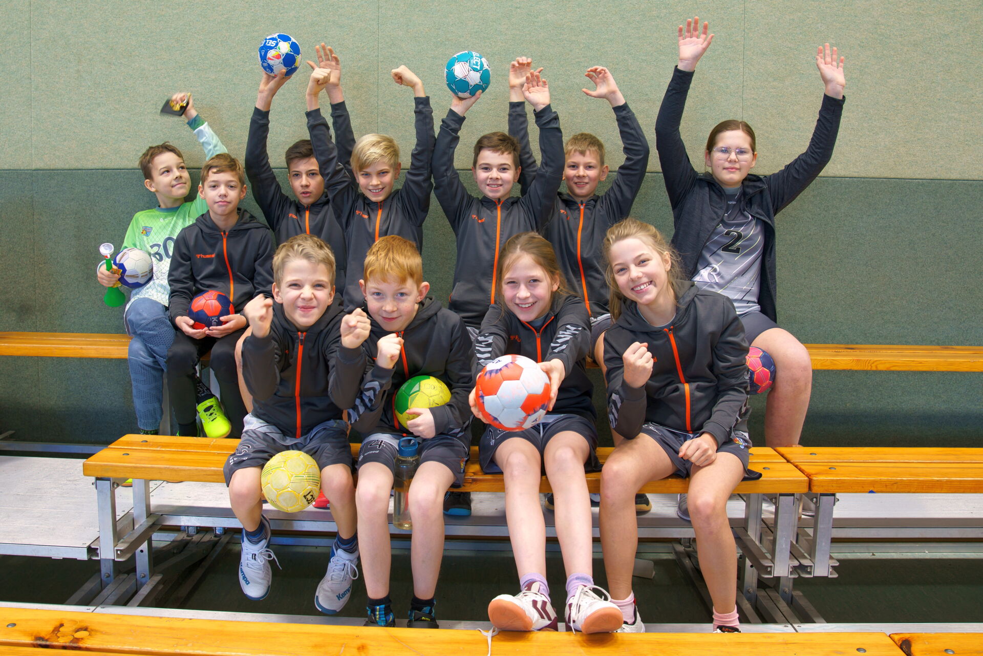 Eine Jugendhandballmannschaft der HSG Coswig sitzt jubelnd mit Bällen auf einer Bank in einer Sporthalle.