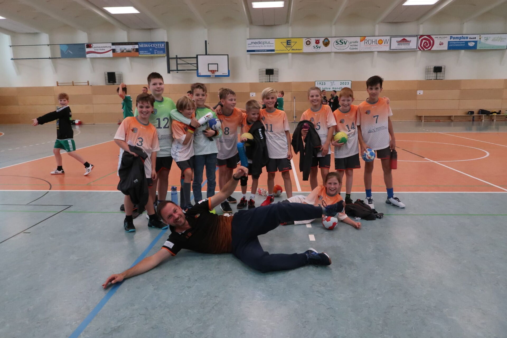 Eine Jugendhandballmannschaft der HSG Coswig posiert mit einem Trainer, der auf dem Boden liegt, in einer Sporthalle für ein Gruppenfoto.