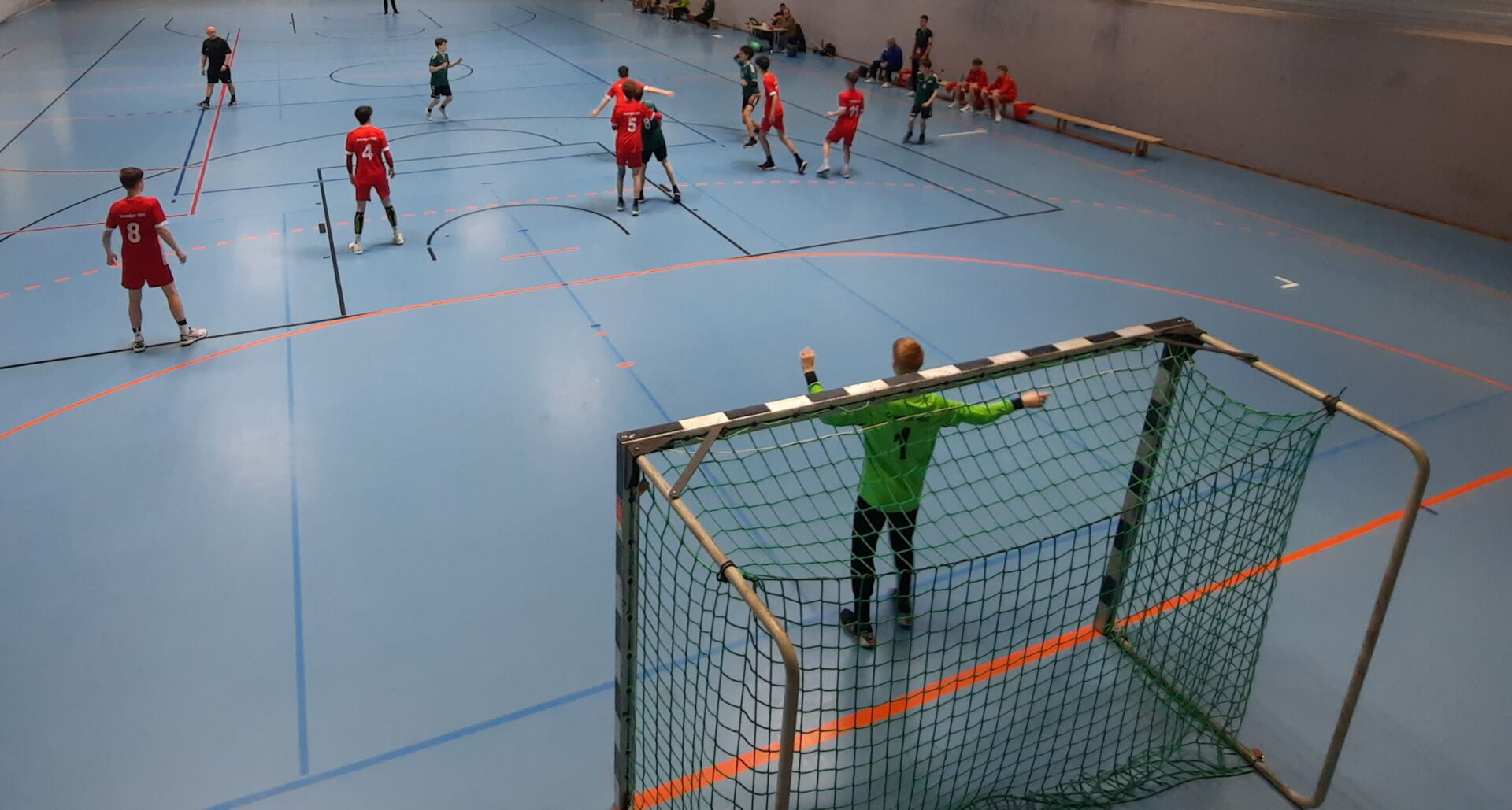Handballspiel in einer Halle. Spieler in roten und grünen Trikots bewegen sich auf dem blauen Feld. Ein Torwart in Grün steht im Vordergrund vor dem Tor.