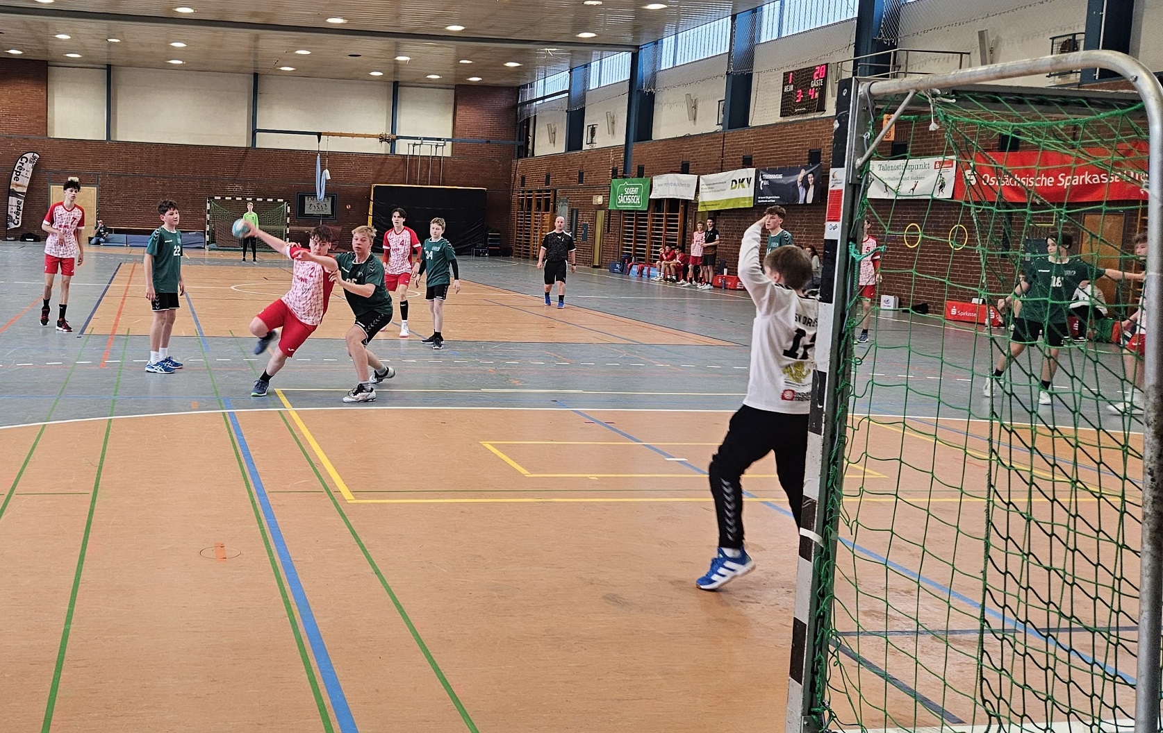 Handballspiel der HSG Coswig in einer Halle. Spieler in roten und grünen Trikots in Bewegung, ein Torwart springt. Zuschauer im Hintergrund.