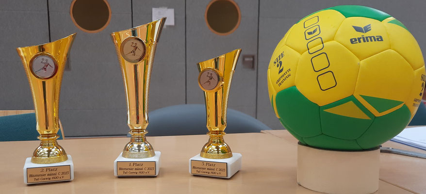 Drei goldene Handball-Pokale mit Plaketten für den 1., 2. und 3. Platz stehen neben einem gelb-grünen Handball der Marke Erima.