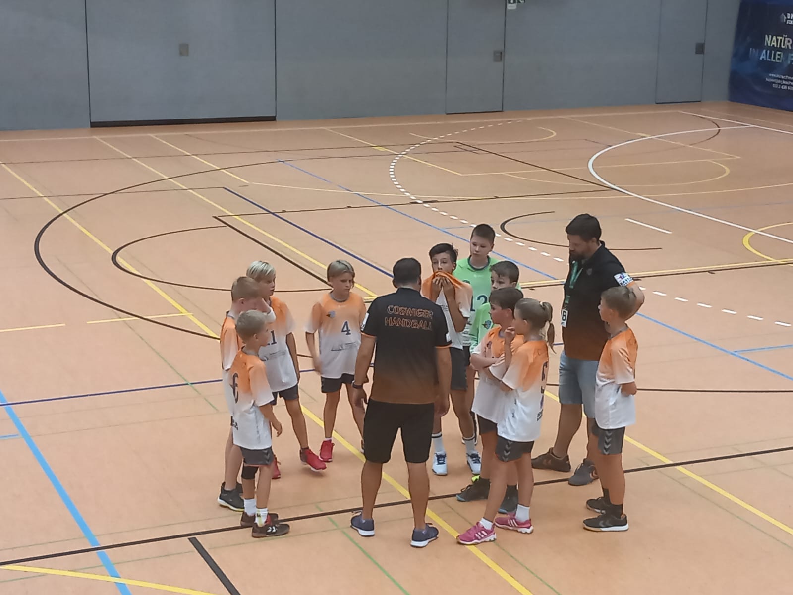 Eine Gruppe junger Handballspieler der HSG Coswig steht in einer Sporthalle im Kreis um zwei Trainer und hört aufmerksam zu.