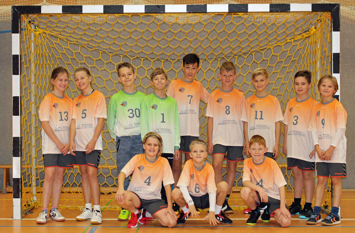 Teamfoto D-Jugend in orangenen-weißen Trikots mit grauer Hose vor einem Handballtor