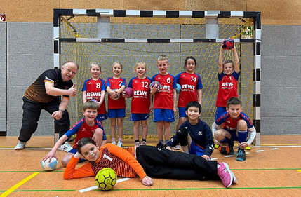Ein Handball-team der HSG Coswig posiert mit Trainer vor einem Handballtor in einer Sporthalle. Alle lächeln in die Kamera.