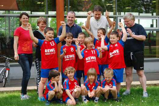 Eine Jugendhandballmannschaft der HSG Coswig mit Trainern jubelt mit erhobenen Fäusten auf einer grünen Wiese.