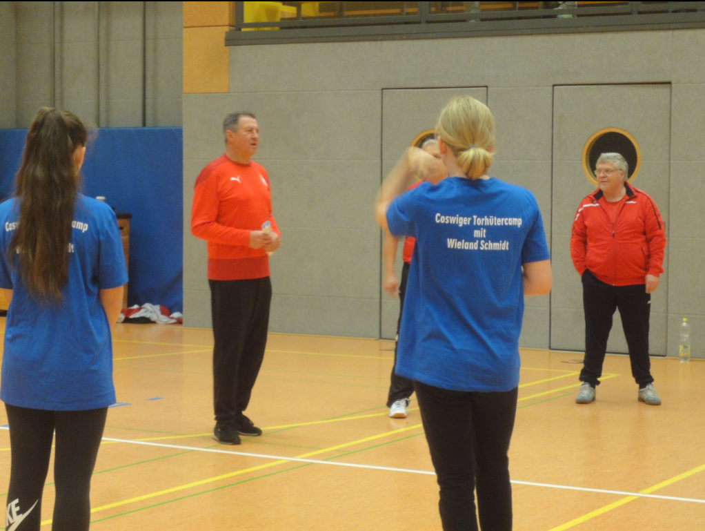 Handball-Torhütercamp der Coswiger Handballspielgemeinschaft in einer Halle. Erwachsene und Jugendliche in Trainingskleidung stehen auf dem Spielfeld.
