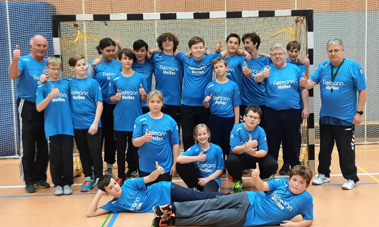 Eine Handballmannschaft der HSG Coswig mit Trainern posiert vor einem Handballtor und zeigt die Daumen nach oben.
